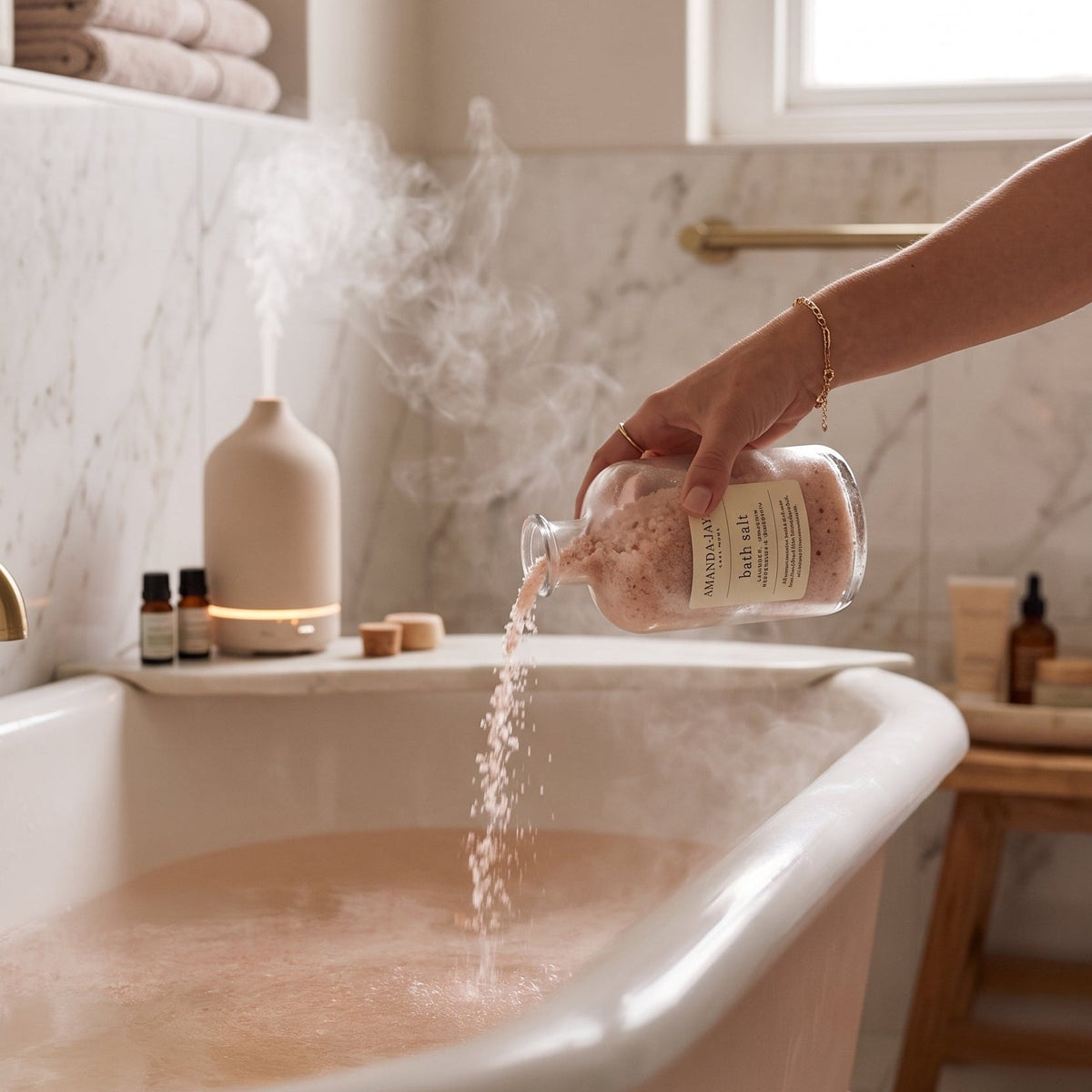 A hand pours pink bath salts from a clear bottle into a bathtub, creating a steaming bath. The scene is set in a bright, modern bathroom with marble tiles, folded towels, and a diffuser.