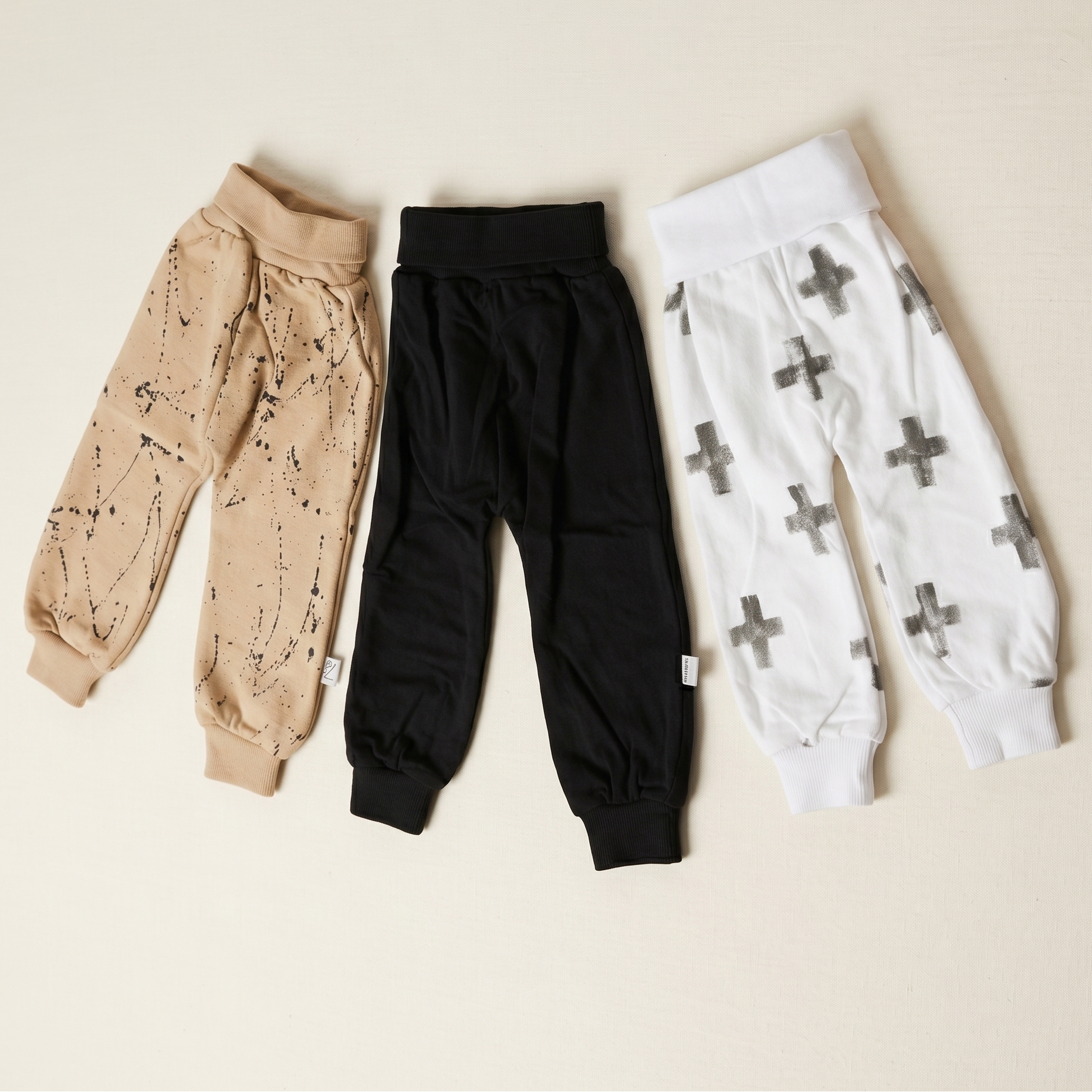 Three pairs of baby leggings in beige, black, and white with patterns on a beige background.