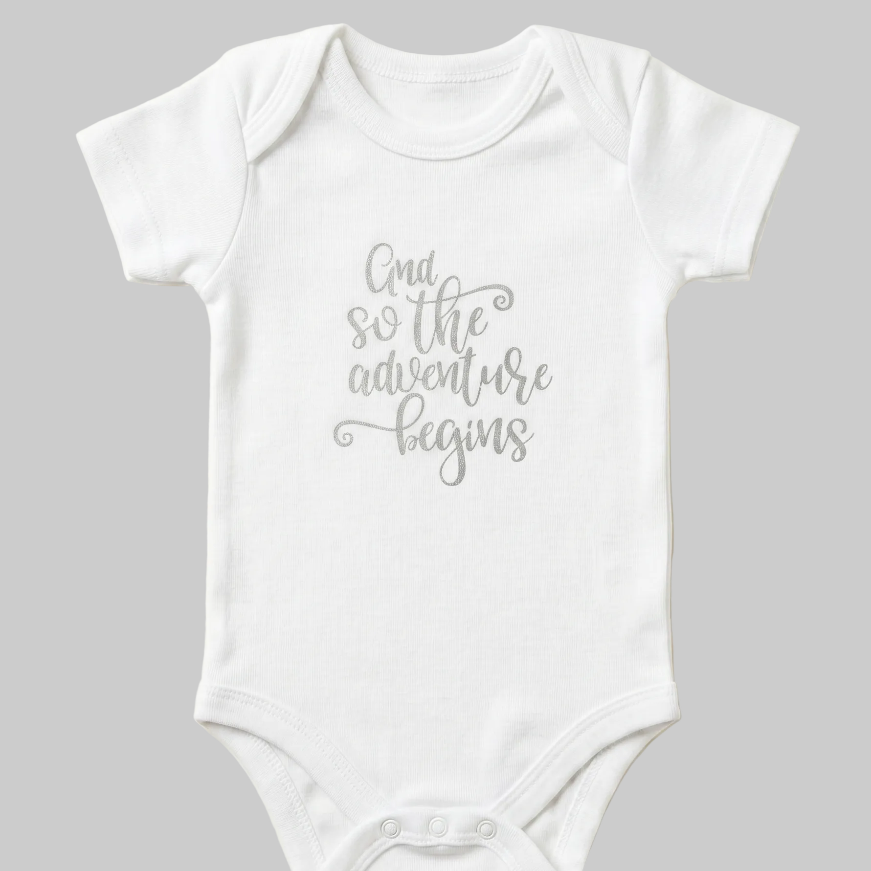 White baby onesie with 'And so the adventure begins' text on a beige background