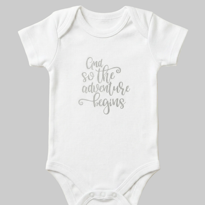 White baby onesie with 'And so the adventure begins' text on a beige background