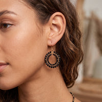 A close-up of a woman wearing a dangling hoop earring with black beaded detailing and a gold-tone metal frame, shown against a softly blurred indoor background.