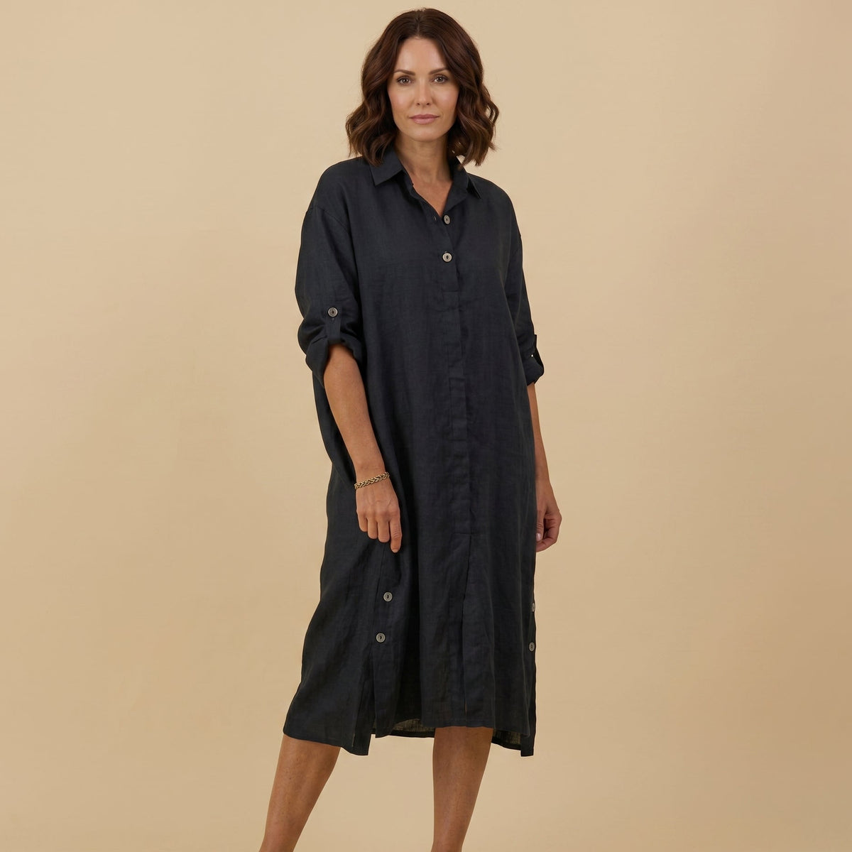 A woman modeling a black linen shirt dress with rolled-up sleeves and button details, standing against a solid beige studio background.