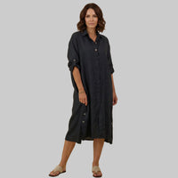 A woman modeling a dark charcoal gray linen shirt dress with rolled-up sleeves and button details, standing against a solid beige studio background.