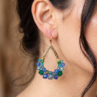 A close-up of a person's ear wearing a dangling earring. The earring has a gold-tone hook and braided top, with a circular lower section made of faceted beads in blue, green, and clear tones.