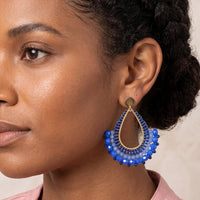 A close-up of a woman wearing a large, ornate teardrop-shaped earring with blue beaded details and gold-toned metal accents, paired with a light pink top against a neutral beige background.