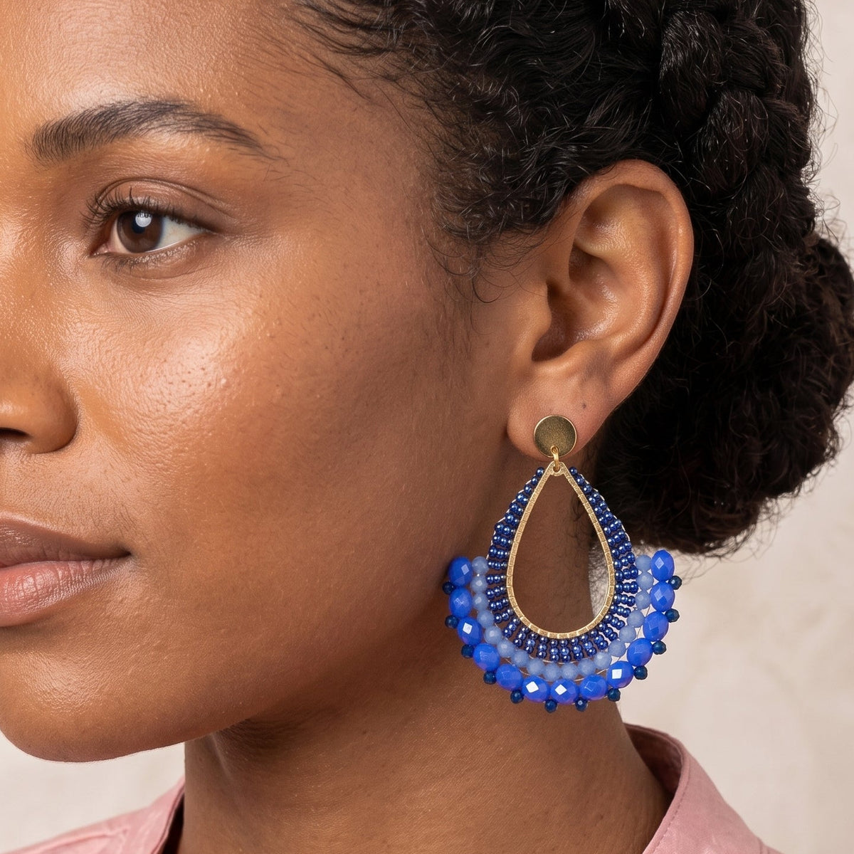 A close-up of a woman wearing a large, ornate teardrop-shaped earring with blue beaded details and gold-toned metal accents, paired with a light pink top against a neutral beige background.