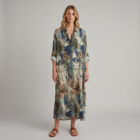 A woman modeling a long-sleeve button-up shirt dress with a tropical leaf print in shades of blue, beige, and brown, paired with brown leather slide sandals, against a neutral gray studio background.