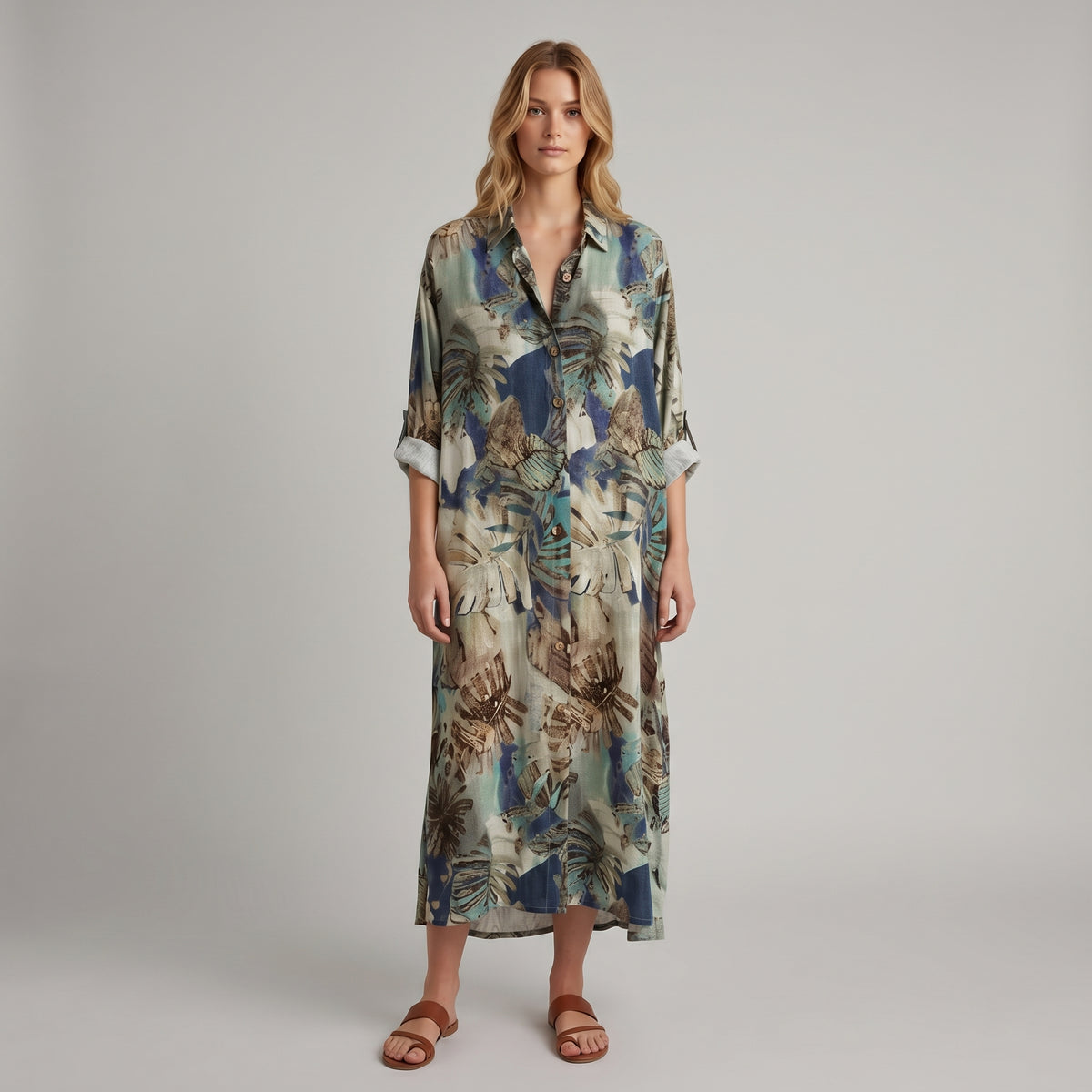A woman modeling a long-sleeve button-up shirt dress with a tropical leaf print in shades of blue, beige, and brown, paired with brown leather slide sandals, against a neutral gray studio background.