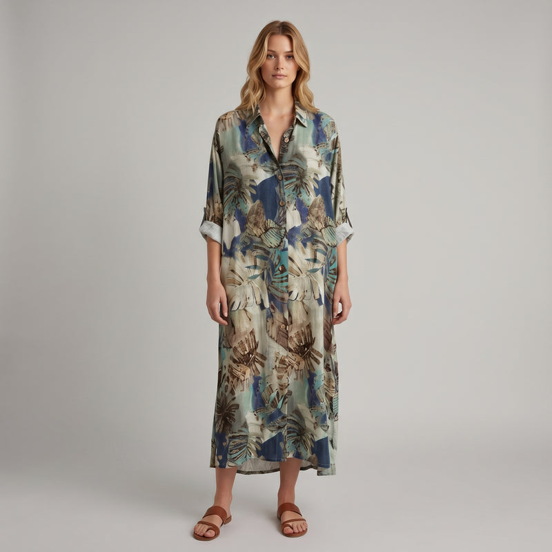 A woman modeling a long-sleeve button-up shirt dress with a tropical leaf print in shades of blue, beige, and brown, paired with brown leather slide sandals, against a neutral gray studio background.