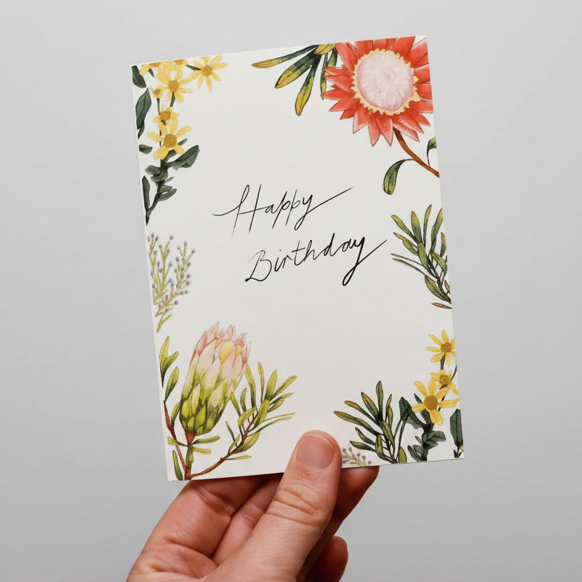 A hand holds a square birthday card with a watercolor floral design and the words 'Happy Birthday' in black script. The card is surrounded by other illustrated cards on a light stone surface.