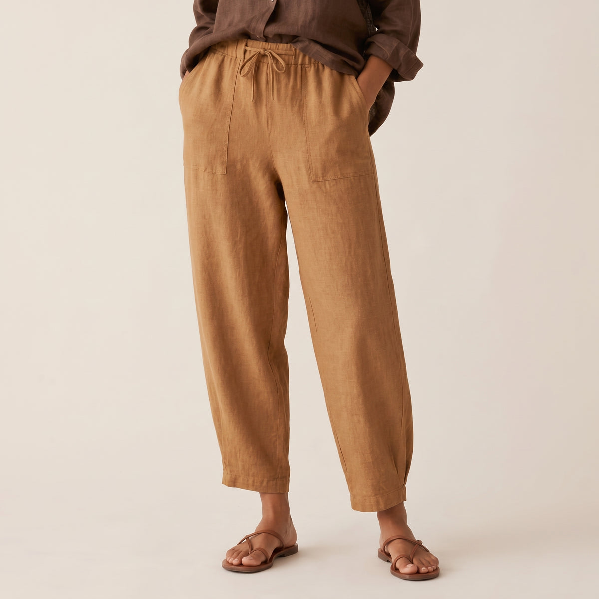 A person wearing loose, wide-leg tan linen pants with a drawstring waist and side pockets, paired with a dark brown shirt and brown sandals, against a plain beige studio background.