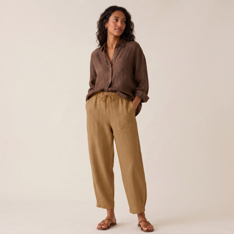 A woman modeling a brown button-up linen shirt and tan linen drawstring pants against a warm off-white studio background.