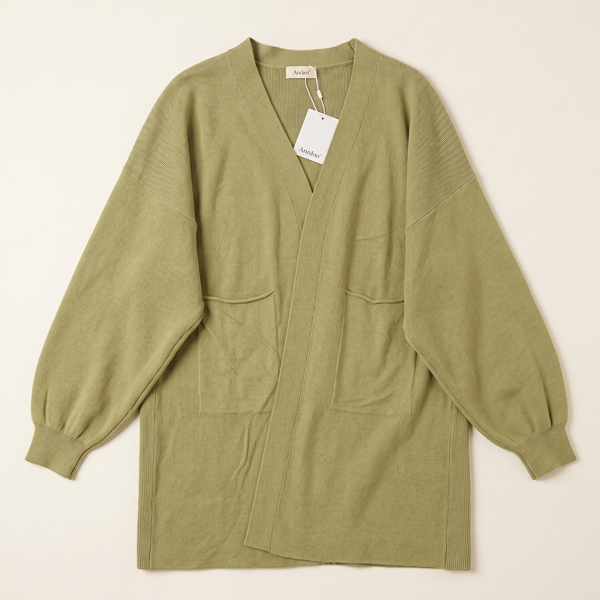 An olive green knit cardigan sweater with long balloon sleeves, a split V-neck, and two front patch pockets, laid flat on a light beige background with a white tag attached.
