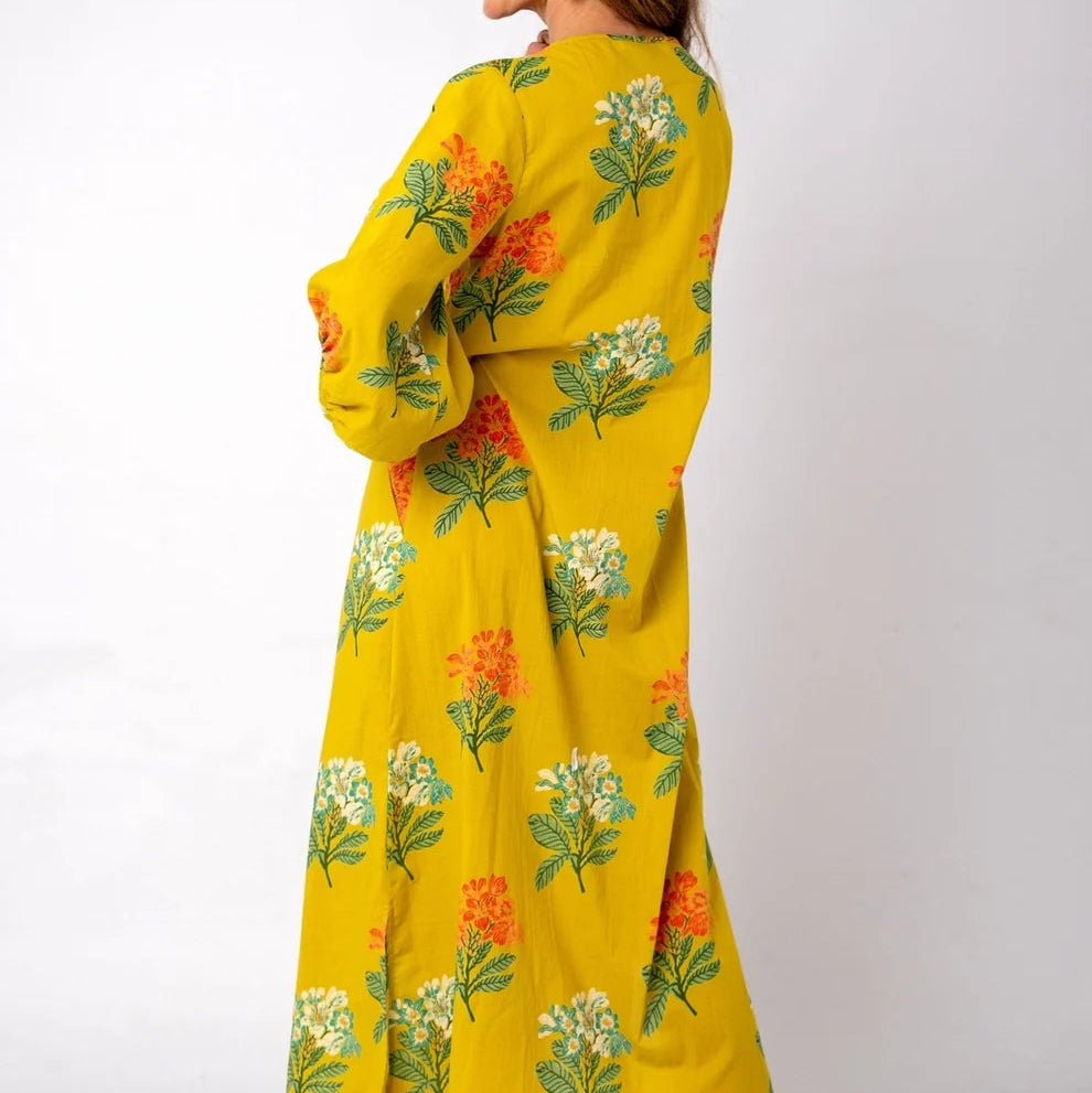 A woman modeling a long yellow dress with a green, orange, and white floral print, shown from a side-back angle against a plain white studio background.