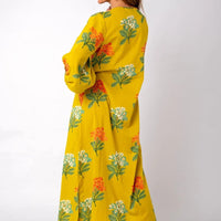 A woman modeling a long yellow dress with a green, orange, and white floral print, shown from a side-back angle against a plain white studio background.