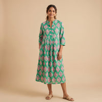 A woman modeling a green midi dress with a pink floral ikat pattern, standing against a solid beige studio background.