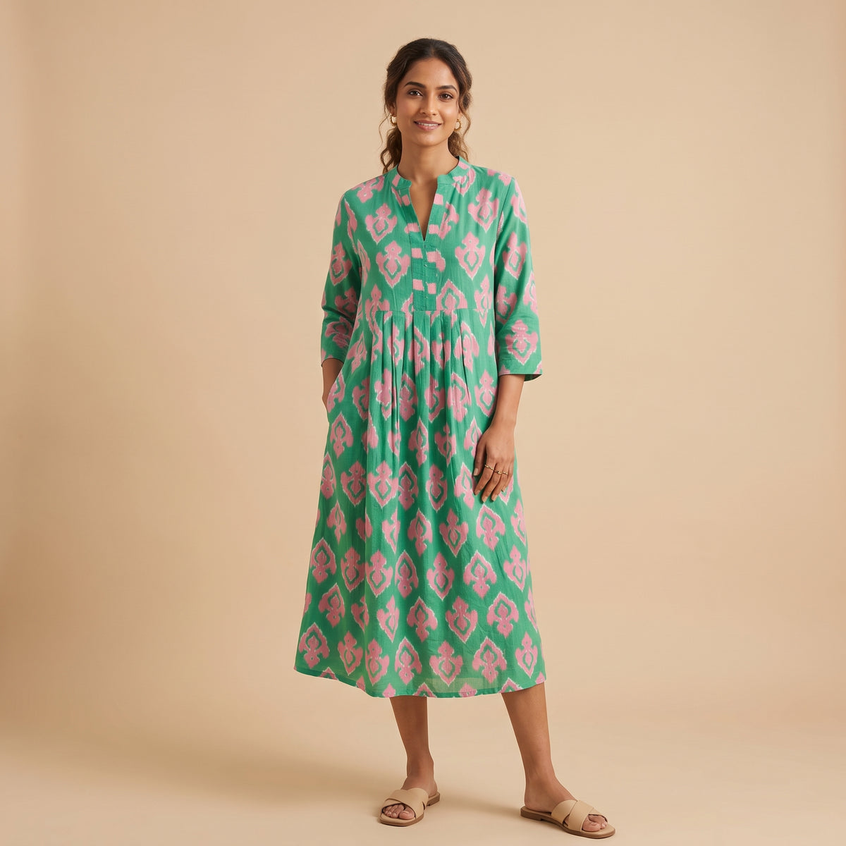 A woman modeling a green midi dress with a pink floral ikat pattern, standing against a solid beige studio background.
