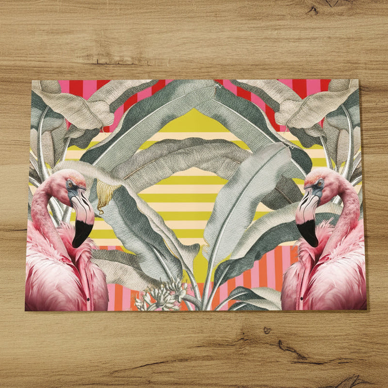 A rectangular placemat with a vibrant tropical design featuring pink flamingos, large green banana leaves, and striped pink, red, and yellow backgrounds, displayed on a light wood surface next to a seed-covered snack ball.