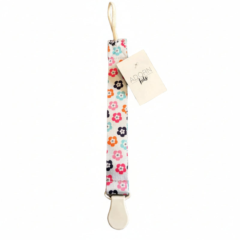 Floral-patterned pacifier clip with a 'Adorn Kids' tag on a brown surface