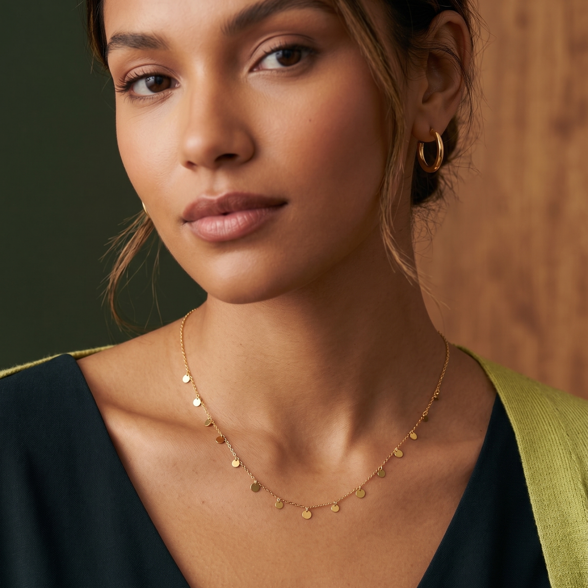 A close-up of a woman wearing a delicate gold chain necklace with small flat disc charms, styled with a dark green V-neck top and a light green shoulder accent.