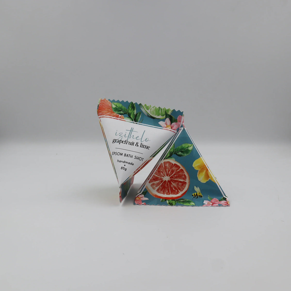Two triangular bath salts packets with a colorful citrus and floral design, standing upright on a light gray background. The front packet shows the brand name 'isothelo' and the scent 'grapefruit & lime'.
