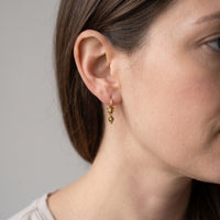 A close-up of a woman's ear wearing a delicate gold hoop earring with two small dangling green beads, shown against a plain light gray background.