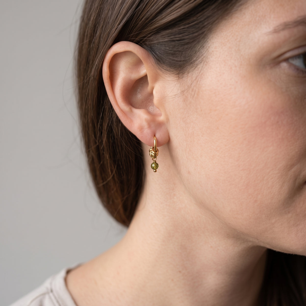 A close-up of a woman's ear wearing a delicate gold hoop earring with two small dangling green beads, shown against a plain light gray background.
