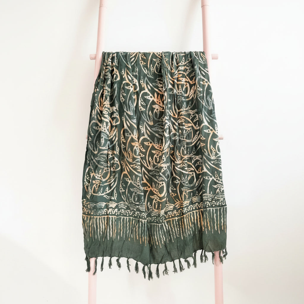 Green and gold patterned scarf with fringes on a wooden hanger against a neutral background
