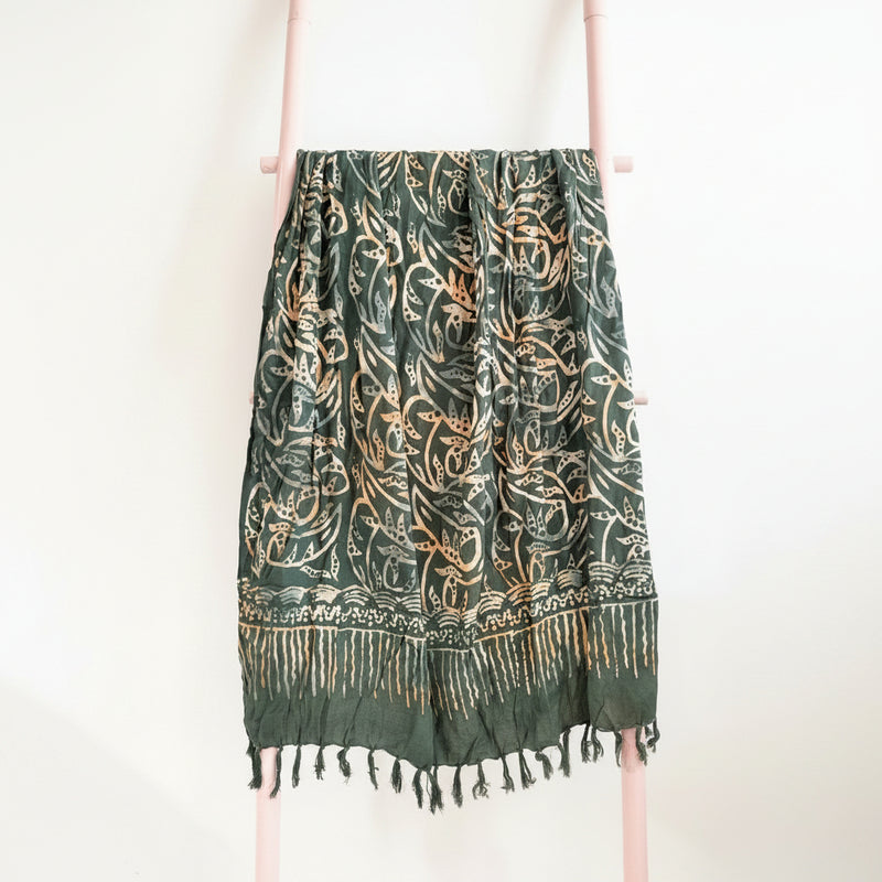 Green and gold patterned scarf with fringes on a wooden hanger against a neutral background