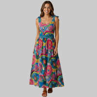 A woman modeling a sleeveless maxi dress with a vibrant, multicolored psychedelic pattern featuring pink, blue, yellow, and green. The dress has tie-shoulder straps, a smocked bodice, and side pockets, shown against a solid beige studio background.