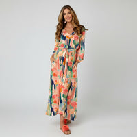 A woman modeling a long-sleeve maxi dress with a colorful abstract floral print, featuring a tie waist and side pockets, set against a plain white studio background.