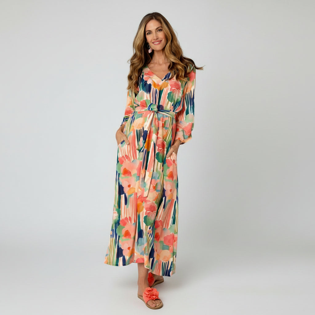 A woman modeling a long-sleeve maxi dress with a colorful abstract floral print, featuring a tie waist and side pockets, set against a plain white studio background.