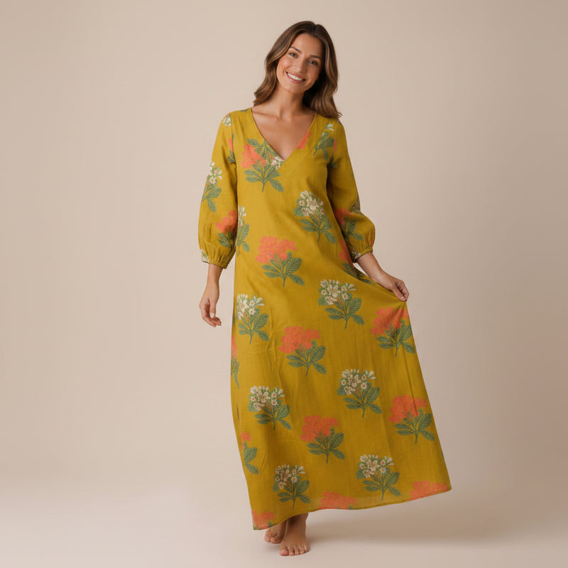 A woman modeling a long-sleeved mustard yellow maxi dress with a green and orange floral print, standing against a solid beige background.