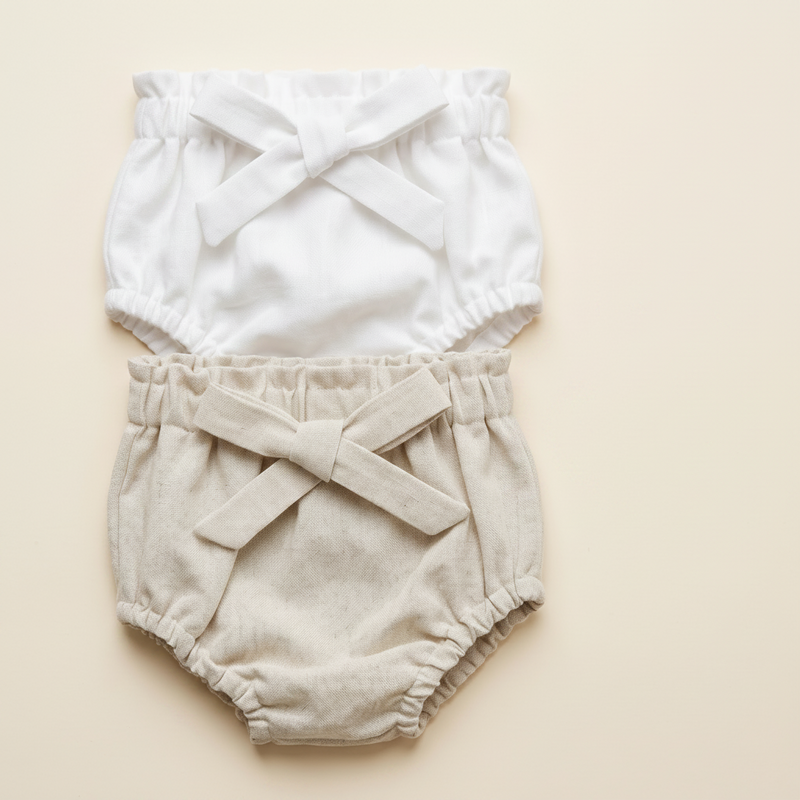 Baby bloomers with criss-cross straps on a beige background with a brand tag.