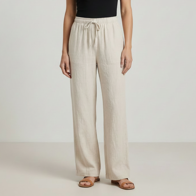 A model wearing light beige wide-leg linen pants with an elastic drawstring waist, paired with a black top and brown leather slide sandals, set against a neutral studio background.