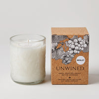A white wax candle in a clear textured glass vessel stands beside its kraft paper box. The box features an illustration of grapes and the text 'UNWINED' and 'MERLOT'.