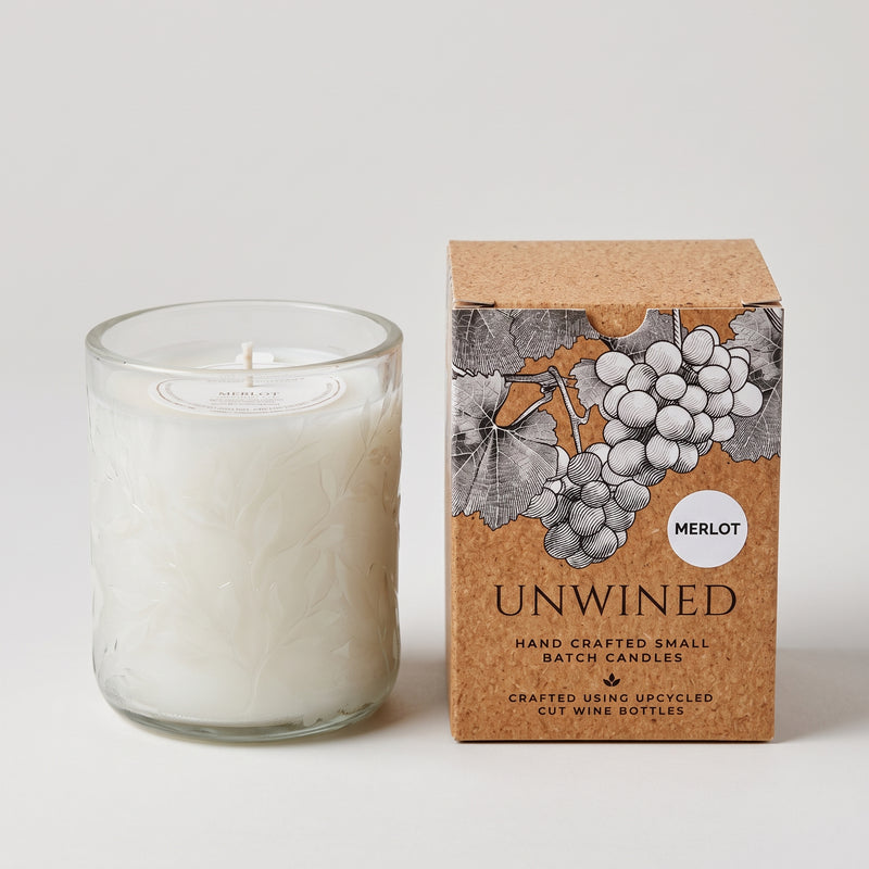 A white wax candle in a clear textured glass vessel stands beside its kraft paper box. The box features an illustration of grapes and the text 'UNWINED' and 'MERLOT'.