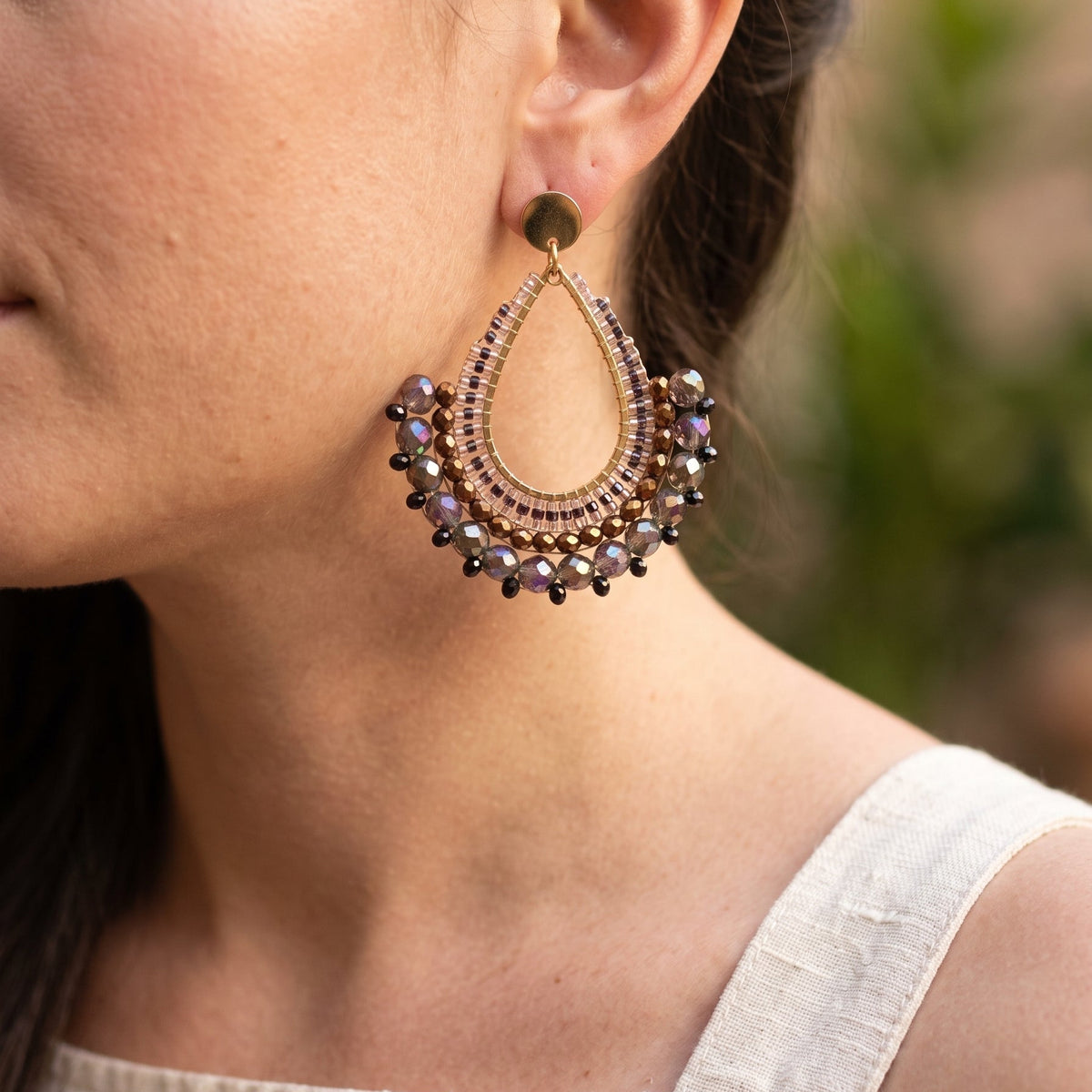 A close-up side profile of a woman wearing a large teardrop-shaped statement earring. The earring features a bronze-toned metal frame, intricate beadwork, and dangling dark purple and black beads.