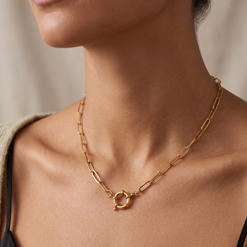 A close-up of a model wearing a gold paperclip chain necklace with a circular ring clasp, styled with a black camisole top.