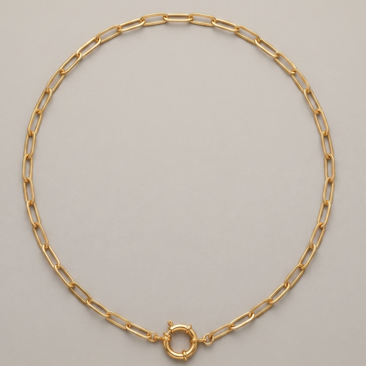 A gold paperclip chain necklace with a small circular ring clasp, displayed on a neutral light gray background.
