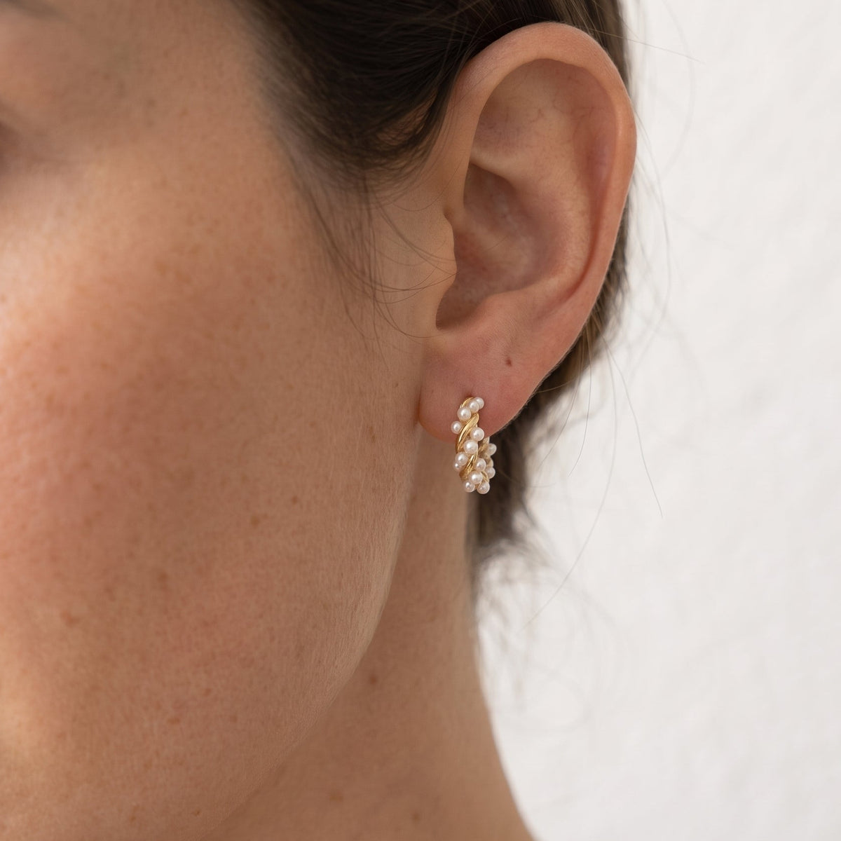A close-up of a woman's ear wearing a small gold earring adorned with clustered white pearls, set against a plain off-white wall.