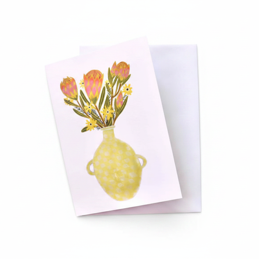 A square greeting card with a watercolor-style illustration of a yellow vase holding pink and yellow flowers, placed on top of a white envelope over a rustic whitewashed wooden surface.