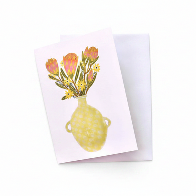 A square greeting card with a watercolor-style illustration of a yellow vase holding pink and yellow flowers, placed on top of a white envelope over a rustic whitewashed wooden surface.