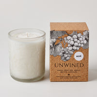 A white scented candle in a clear glass jar sits beside its kraft paper box. The box features a black and white illustration of grape clusters and the text 'UNWINED' and 'ROSÉ'.