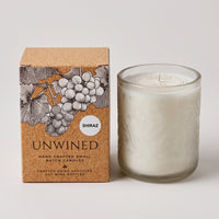 A white scented candle in a clear glass jar stands beside its kraft paper box, which features an illustration of grapes and the text 'UNWIRED' and 'SHIRAZ'.