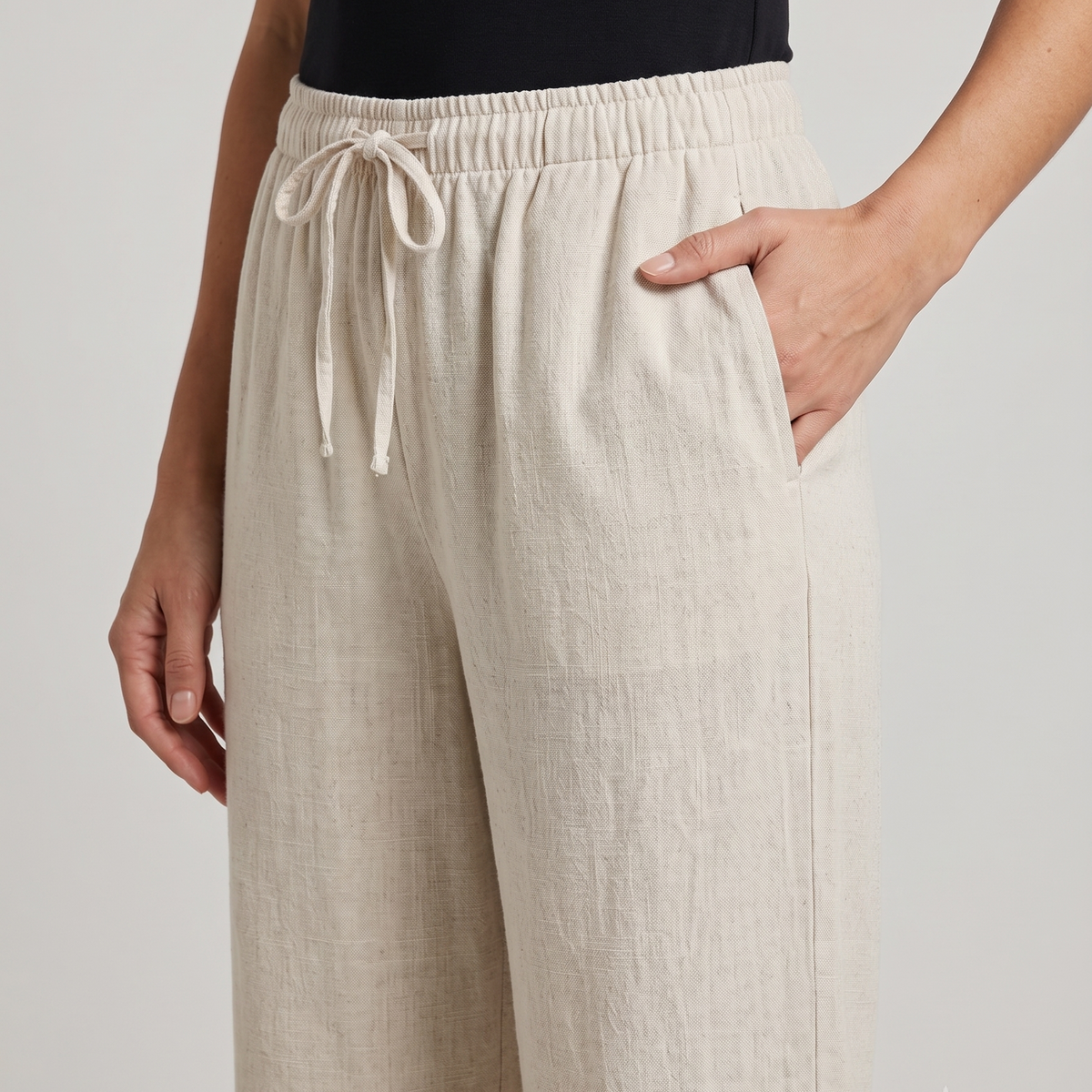 A close-up of a person wearing light beige wide-leg drawstring pants with side pockets, paired with a black top, against a plain light gray studio background.