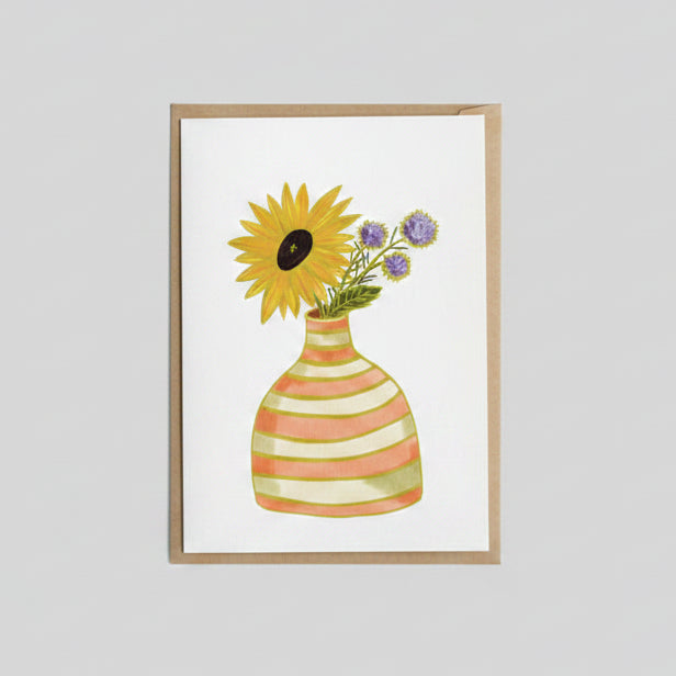 A square greeting card with a watercolor-style illustration of a yellow sunflower and purple flowers in a striped vase, placed on a brown wooden surface with another card partially visible at the right edge.