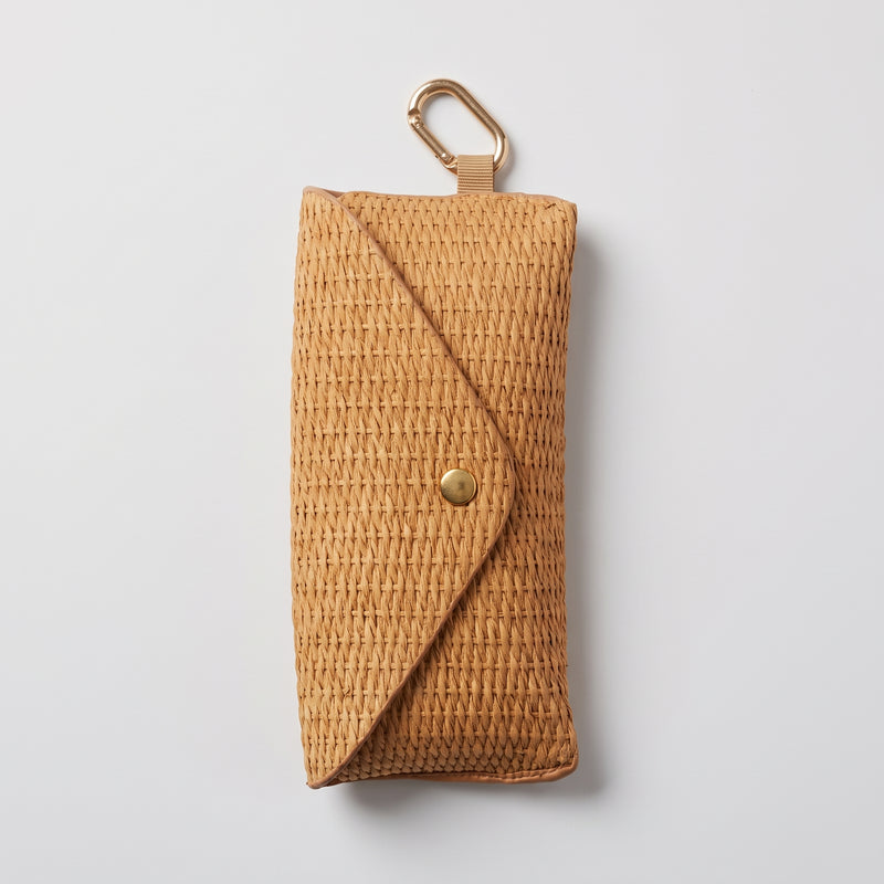 A natural woven straw pouch with a diagonal flap closure and a brass snap button, featuring a gold carabiner clip at the top, shown on a light gray background.