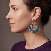 A woman in profile wearing a large teardrop-shaped dangle earring with blue beads and gold-toned metal details, set against a neutral brown studio background.