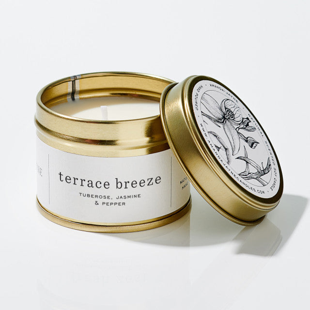 A gold metal candle tin with the lid off, showing a white wax candle and a white label that reads 'terrace breeze' with scent notes 'TUBEROSE, JASMINE & PEPPER.' The brand name 'MORNING GLOW' is visible on the lid illustration.